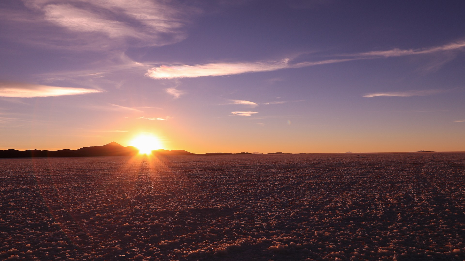 Sunrise over a flat desert landscape with distant mountains silhouetted against the horizon