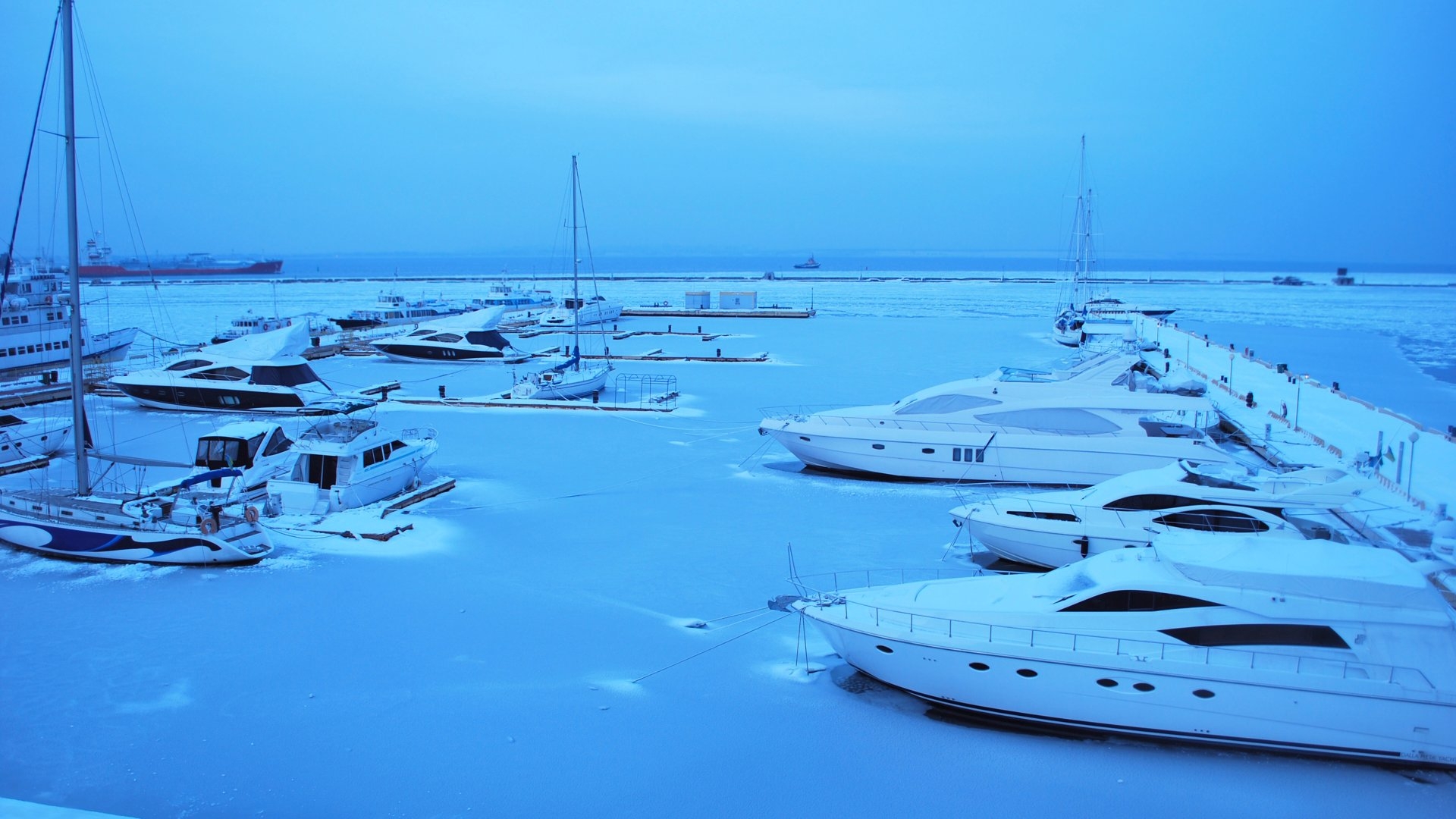 Luxury yachts and sailboats docked at a marina during winter conditions with icy water and snow-covered piers