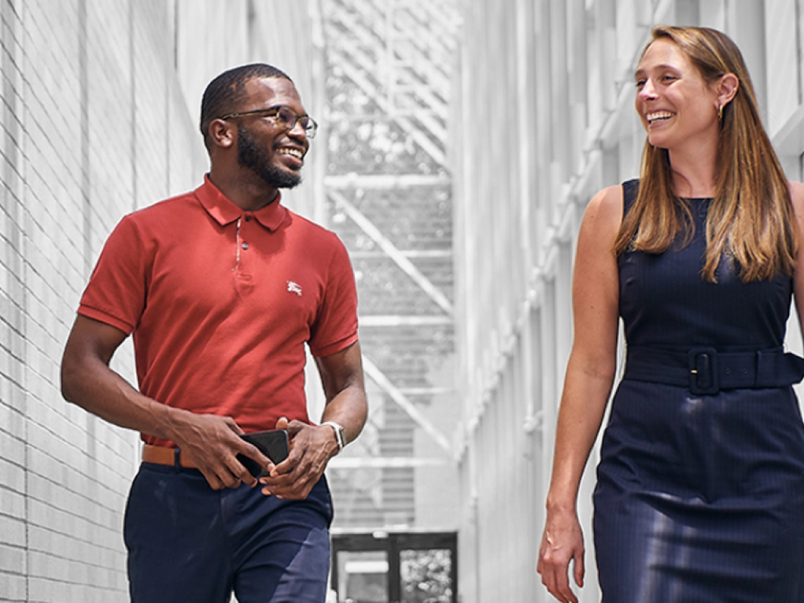 a black male in a red cummins polo and white female in a navy dress smile and talk while walking down a white hallway